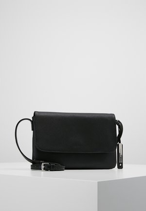 Sac bandoulière en cuir noir avec fermeture à rabat, sangle réglable et détail d'étiquette métallique sur une surface blanche.