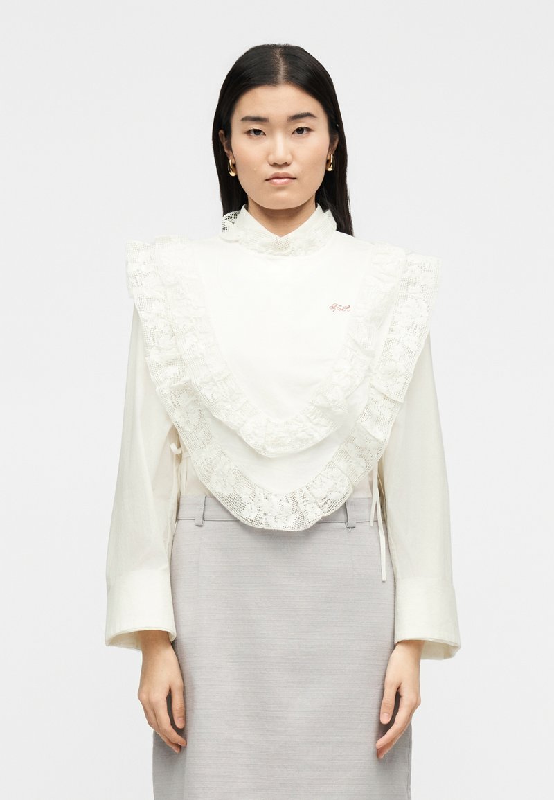 Γυναίκα με ίσια σκούρα μαλλιά που φοράει λευκή μπλούζα με στρώσεις δαντέλας στο μπροστινό μέρος και γκρι φούστα, στέκεται μπροστά σε απλό φόντο.