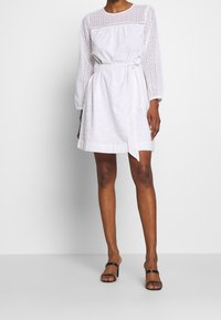 Vestido blanco con manga larga, tejido de ojales, cuerpo ajustado, cinturón en la cintura y longitud por encima de la rodilla; combinado con sandalias negras de tiras.