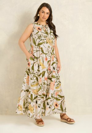 Femme portant une robe maxi sans manches à étages avec un motif floral et des sandales compensées marron, debout devant un fond beige uni.
