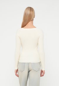 Ριχτό μπλου�ζάκι με μακριά μανίκια σε εκρού απόχρωση, με στρογγυλή λαιμόκοψη, εφαρμοστή γραμμή και μαλακή υφή, συνδυασμένο με τζιν σε ανοιχτό χρώμα.