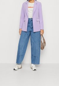 Blazer de lavanda sobre una camisa blanca de rayas con texto rojo, combinado con jeans de pierna ancha azules y zapatillas blancas y marrones. Sostiene un bolso de patrón de serpiente.