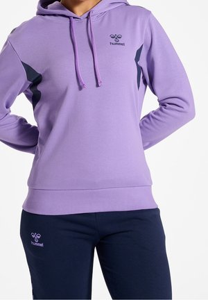 Person iført en lilla hummel-hættetrøje med sorte accenter og navy hummel-bukser, stående med hænderne bag ryggen på en hvid baggrund.