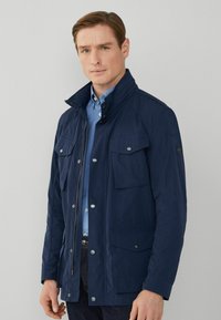 Marinblå jacka med hög krage, framzipper, fyra framfickor och silverfärgade detaljer. Tillverkad av ett slätt, slitstarkt material.