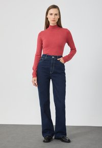 Czerwony dopasowany sweter z golfem w zestawieniu z wysokim stanem ciemnoniebieskich dżinsów. Gładka faktura, wyraźne szwy i klasyczny krój prosty.