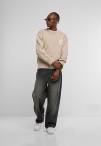 Beiger Strickpullover mit einem gerippten Halsausschnitt und weißem Emblem, kombiniert mit grauen weiten Jeans und weißen Sneakern vor einem neutralen Hintergrund.