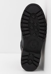 Suela de zapato de goma negra con un diseño de banda de rodadura texturizado y en relieve, presenta estrías para mayor tracción y un logotipo estampado en el centro.