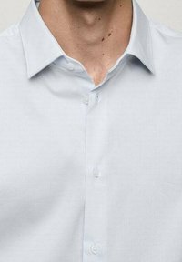 Camisa azul celeste con botones y cuello, confeccionada en una tela lisa. Cuenta con una tapeta frontal con dos botones visibles y una textura sutil.