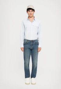Ung mann som ser fremover, iført en beige caps, lyseblå skjorte med knapper, blå jeans og hvite joggesko mot en enkel hvit bakgrunn.
