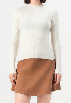 Vrouw draagt een strakke witte geribbelde trui met lange mouwen en een bruine corduroy A-lijn minirok tegen een witte achtergrond.