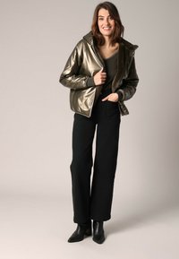 Doudoune métallisée vert olive avec capuche, dotée d'un col en fausse fourrure et de poignets côtelés. Portée par-dessus un haut sombre et un pantalon large noir.