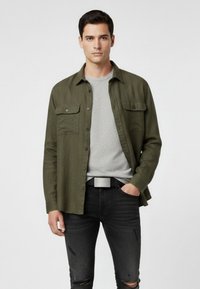 Olive green button-up shirt with two chest pockets over a light gray crew neck t-shirt, paired with black distressed jeans and a silver belt.