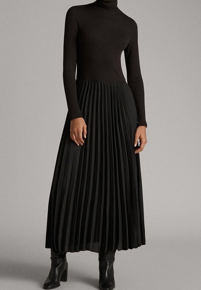 Femme portant un haut à col roulé noir à manches longues et une jupe longue plissée noire, associée à des bottines à talons noires.