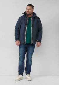 Marineblaue Jacke mit hohem Kragen, Reißverschluss und Taschen, übereinander getragen mit einem grünen Shirt und einem karierten Hemd. Getragen mit dunklen Jeans und Turnschuhen.