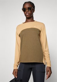 Lange mouwen shirt in beige en olijfgroen, met een ronde hals en textuurstof. Gecombineerd met zwarte legging en een zwarte tas.