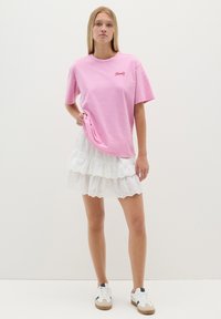 Camiseta de algodón rosa de manga corta con un pequeño logo, combinada con una falda blanca de volantes con ojales en capas. Zapatillas blancas completan el conjunto.