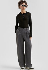 Schwarzes geripptes Langarm-Polohemd mit Kragen, graue weit geschnittene Hose und schwarz-weiße Sneaker. Weiße quadratische Sonnenbrille als Akzent.