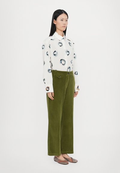 Camisa blanca de manga larga con patrón circular azul, combinada con pantalones de pana verde de pierna ancha y manoletinas a rayas multicolores.