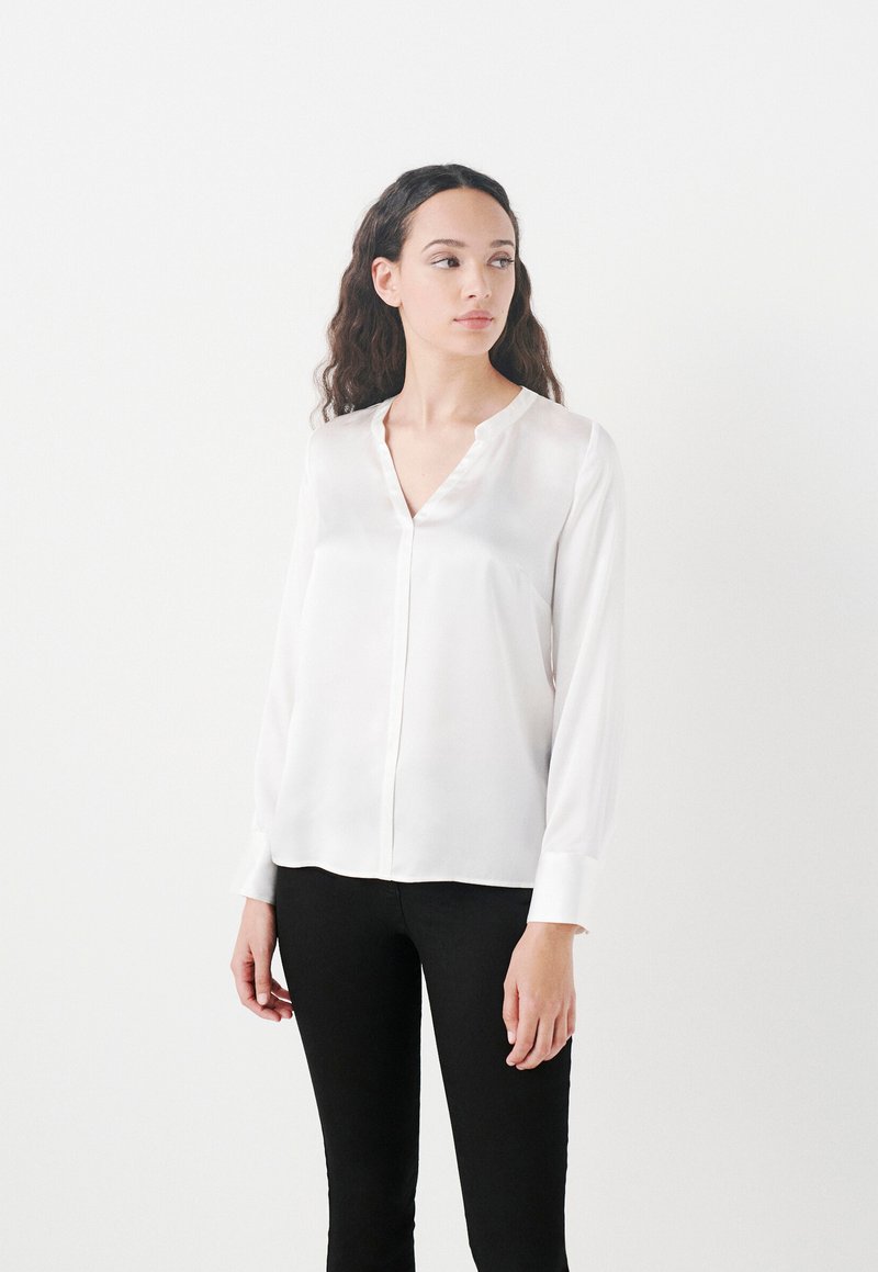 Blusa blanca de manga larga hecha de tejido suave, con escote en V, un brillo sutil y puños abotonados, combinada con pantalones ajustados oscuros.