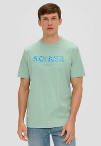 Mintgrünes Baumwoll-T-Shirt mit einem blauen Blumenprint-Logo "S. OLIVER" auf der Brust und einem runden Halsausschnitt, getragen mit blauen Jeans.