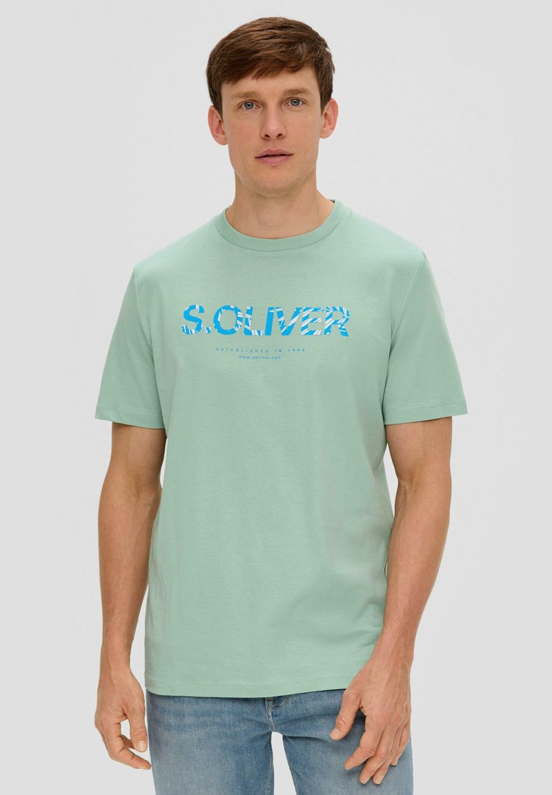 Mintgrünes Baumwoll-T-Shirt mit einem blauen Blumenprint-Logo "S. OLIVER" auf der Brust und einem runden Halsausschnitt, getragen mit blauen Jeans.