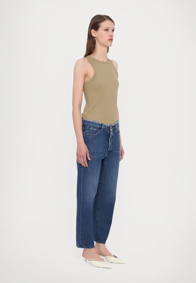 Khaki sleeveless top with a round neckline, paired with loose-fit dark blue denim jeans and white high-heeled mules. Simple design and texture.