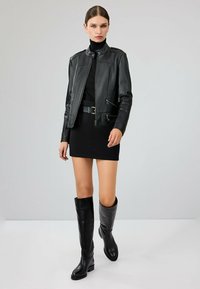 Zwarte leren motorjack met een getailleerd ontwerp, ritsdetails, hoge kraag, gecombineerd met een zwarte minidress en zwarte kniehoogte laarzen.