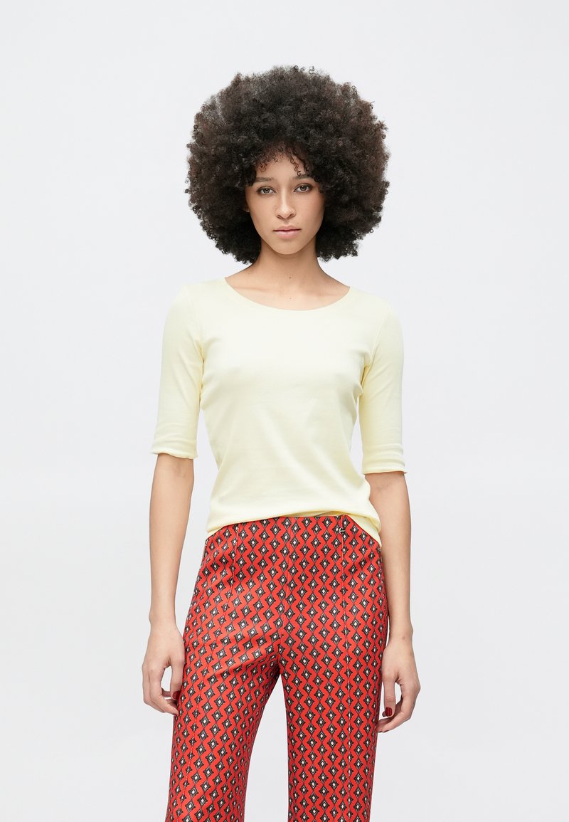 Svetlo rumena oprijeta majica z okrolim izrezom in rokavi do komolcev, kombinirana z rdečimi širokimi hlačami z geometrijskim vzorcem.