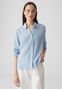 Camicia azzurro chiaro, con bottoni, realizzata in tessuto testurizzato, dotata di colletto, maniche arrotolate e bottoni bianchi. Indossata con pantaloni chiari.