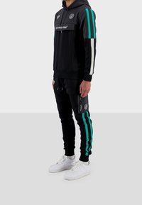 Conjunto de sudadera negra con capucha y pantalones de chándal con rayas en verde azulado y blanco. Presenta un logo en el pecho y un bolsillo con cremallera en los pantalones. Zapatillas blancas.