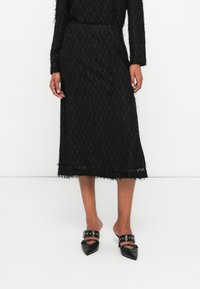 Sort tekstureret midi-nederdel med et diamantmønster, frynse detaljer og en gennemsigtig nederste kant. Kombineret med sorte spidse slip-on sko med stropper.
