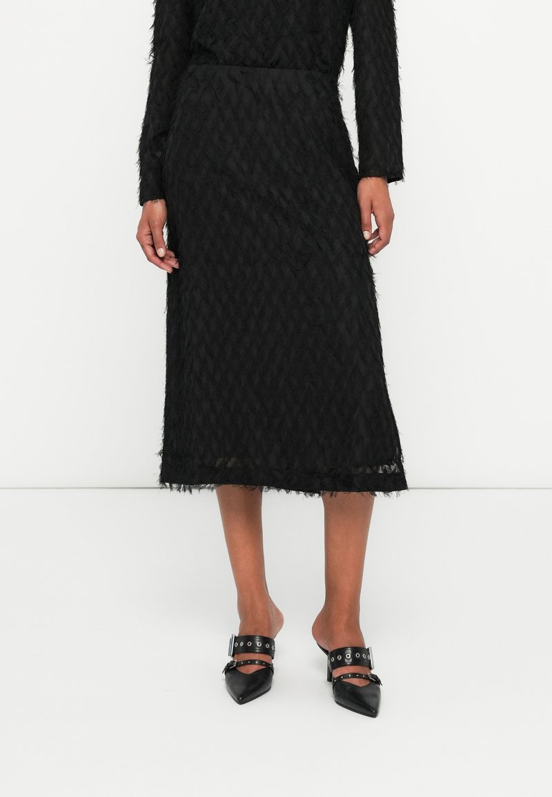 Sort tekstureret midi-nederdel med et diamantmønster, frynse detaljer og en gennemsigtig nederste kant. Kombineret med sorte spidse slip-on sko med stropper.