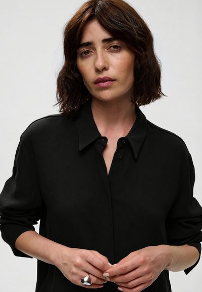 Femme aux cheveux foncés mi-longs portant une chemise noire boutonnée, tenant ses mains jointes devant elle, avec une grosse bague en argent à la main gauche.