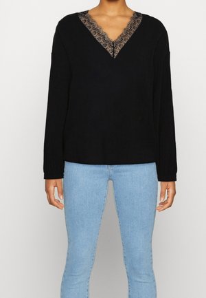 Persoon die een zwarte top met lange mouwen en kant V-hals draagt en lichtblauwe aangesloten jeans, staand tegen een effen achtergrond.