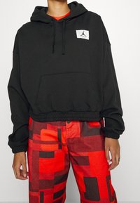 Felpa nera cropped con tasca a marsupio e patch con logo; abbinata a pantaloni rossi con motivo geometrico.
