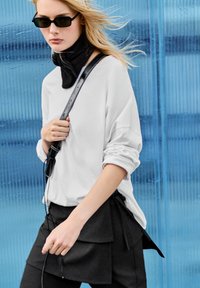White oversized long-sleeve top paired with a black layered skirt. Black sunglasses, black scarf, and a strap bag are also visible.