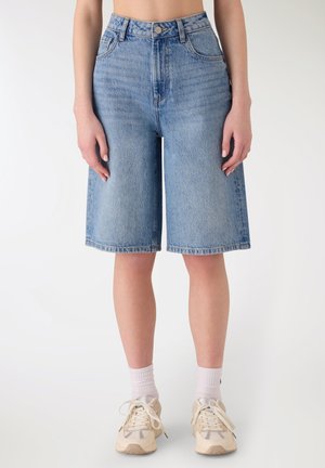 Donna che indossa bermuda in denim azzurro chiaro a vita alta con tasche frontali, in piedi a piedi nudi su uno sfondo bianco.