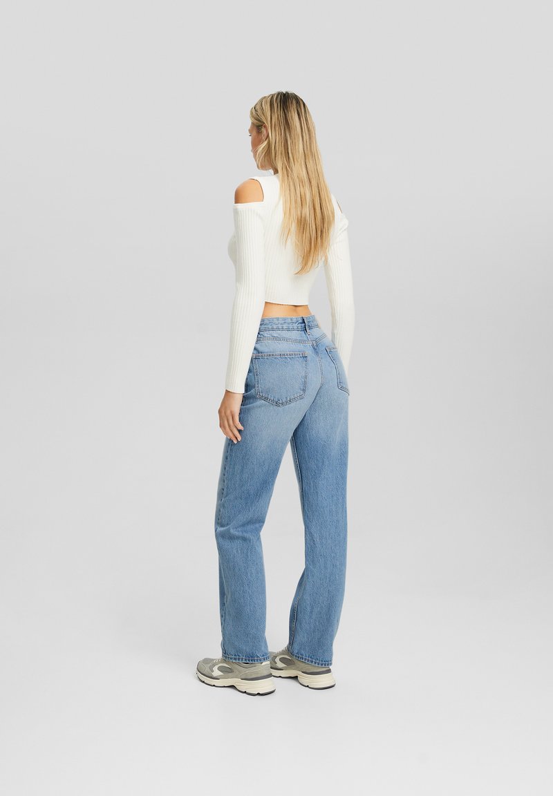 Biały prążkowany crop top z długim rękawem i wyciętymi ramionami, w zestawie z jasnoniebieskimi spodniami o wysokim stanie i prostych nogawkach. Sneakersy dopełniają całość.