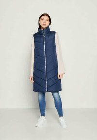 Γυναίκα που στέκεται φορώντας ένα μακρύ ναυτικό γιλέκο με φερμουάρ μπροστά, ανοιχτό μπεζ μακρυμάνικη μπλούζα, μπλε skinny τζιν και λευκά sneakers.