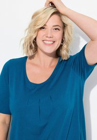 Camicia a maniche corte color teal con scollo rotondo e dettagli di pieghe sottili sul davanti. Tessuto morbido con una consistenza liscia.