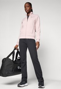 Sweat à zip rose clair, pantalons noirs et baskets noires. Tenant un sac de sport noir avec un logo. Design simple et sportif.