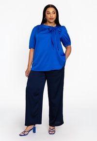 Haut blouson bleu avec un détail de nœud, associé à un pantalon large bleu marine. Le modèle porte des talons bleus à lanières. Tissu lisse avec un léger éclat.