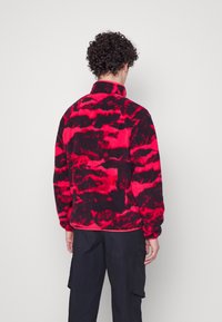 Fleece svetr v červeném a černém tie-dye vzoru s vysokým límcem. Má jemnou strukturu a volný střih, stylizováno s tmavými kalhotami.