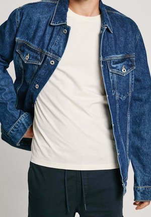 Mand iført en blå denimjakke over en hvid T-shirt og sorte bukser med snor, poserer mod en ensfarvet baggrund.