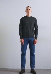 Mees seisab lihtsa heleda seina vastu, seljas must pika varrukaga särk, sinised teksad ja tumedad pruuni värvi saapad, olles suunatud ettepoole.