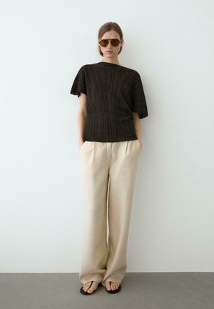 Kvinde iført brun ribbet kortærmet top, beige vidde bukser, sorte sandaler og brune pilot-solbriller, stående mod en hvid væg.