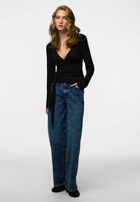 Melns garroku wrap tops ar V-veida izgriezumu un auklas detaļu, kopā ar augstas jostasvietas, platu kāju zili džinsa biksēm. Melni apavi.