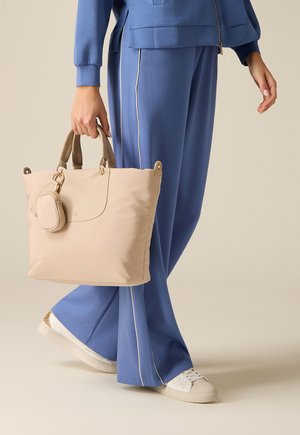 Henkilö sinisissä housuissa ja valkoisissa tennareissa pitää beigeä kangaskassia, jossa on kiinnitetty pieni pussi, seisoo yksinkertaisen taustan edessä.