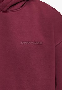 Tissu de sweat à capuche bordeaux avec le texte "DROPSIZE" en relief embossé sur la poitrine près de la couture de l'épaule.