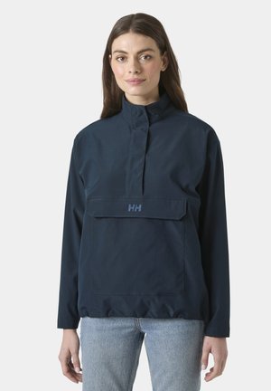 Vrouw draagt een marineblauwe windjack met hoge hals, voorvak met klep en lichtblauwe spijkerbroek, staand tegen een effen lichte achtergrond.